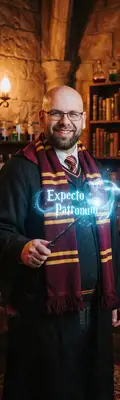 A magical cinematic portrait of the subject as a wizarding school student, wearing dark academy robes with a house-colored scarf, holding a wooden wand casting a glowing blue spell, cozy stone castle common room background with shelves of potions and ancient books, soft warm lighting.