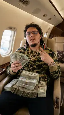 Take the uploaded photo of the person and generate a photorealistic 9х16 image of this person (please make sure to keep facial similarity) in one of the following randomly selected settings:   Leaning against a bright Lamborghini or Rolls-Royce, wearing a tactical vest or puffer jacket, showing off a luxury watch or chain.  Inside a private jet with stacks of cash on their lap, dressed in designer streetwear (like BAPE, Louis Vuitton, or Nike Tech).   Posing in front of a private jet at night, holding fanned-out stacks of hundred-dollar bills, with a confident or nonchalant expression.   Sitting in a luxury car with a starlight ceiling, wearing iced-out chains and sunglasses, mid-conversation or staring coolly ahead.   Standing against an urban graffiti backdrop, throwing up hand gestures while wearing a designer scarf or hoodie, heavy jewelry, and rings.   The final image should have a high-contrast, bold, Instagram-worthy aesthetic. Use realistic lighting and textures. Make sure the background, outfit, and pose reflect confidence, wealth, and a street-smart vibe. Rotate between settings randomly each time this is run.